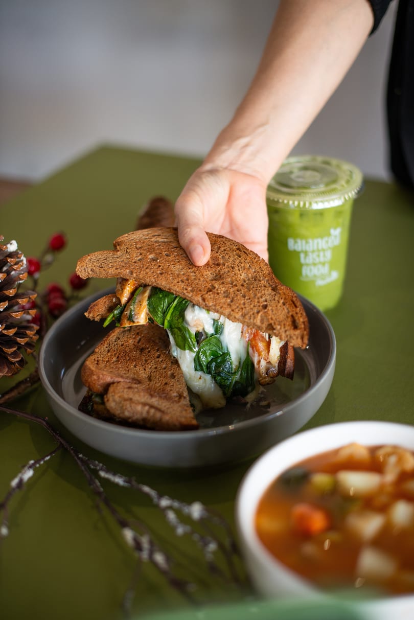 Hand reaching for a toasted sandwich with melted cheese and spinach, next to a green smoothie.