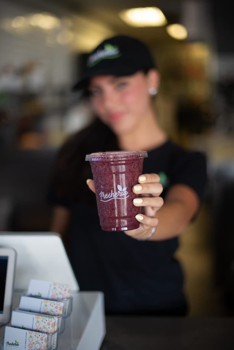 A Fresheria employee in a black cap and shirt holds out a berry smoothie.