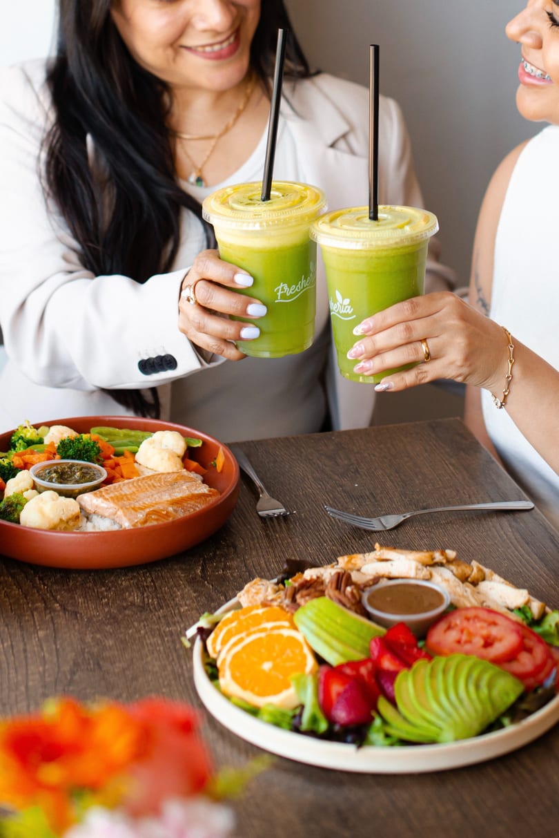 Two women toasting green Fresheria smoothies over plates of salmon and salad.