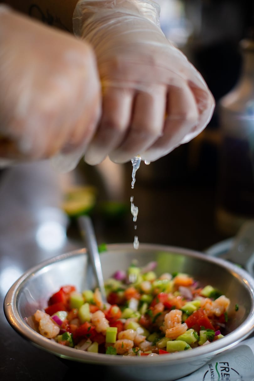 Gloved hands squeeze a lime over a bowl of fresh shrimp ceviche with diced vegetables.